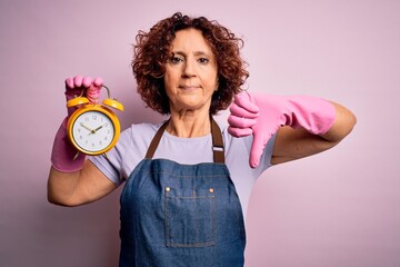 Sticker - Middle age woman cleaning doing housework wearing apron and gloves holding alarm clock with angry face, negative sign showing dislike with thumbs down, rejection concept