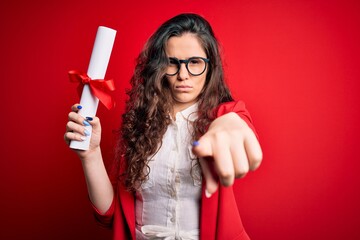 Sticker - Young beautiful woman with curly hair holding university diploma degree over red background pointing with finger to the camera and to you, hand sign, positive and confident gesture from the front