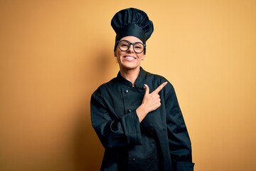 Canvas Print - Young beautiful brunette chef woman wearing cooker uniform and hat over yellow background cheerful with a smile of face pointing with hand and finger up to the side with happy and natural expression