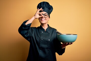 Poster - Young beautiful brunette chef woman wearing cooker uniform and hat holding bowl and whisk with happy face smiling doing ok sign with hand on eye looking through fingers