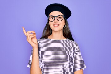 Sticker - Young beautiful brunette woman wearing glasses and french beret over purple background with a big smile on face, pointing with hand and finger to the side looking at the camera.