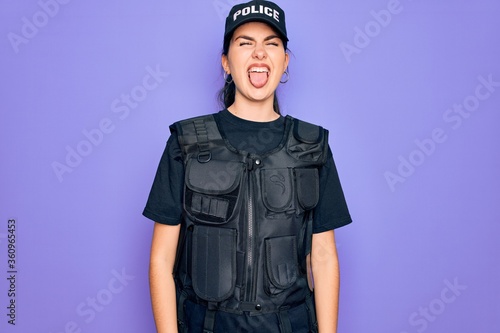 Fotografía Young police woman wearing security bulletproof vest uniform over purple background sticking tongue out happy with funny expression