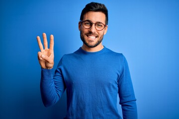 Young handsome man with beard wearing casual sweater and glasses over blue background showing and pointing up with fingers number three while smiling confident and happy.