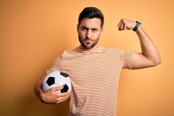 Sticker - Handsome player man with beard playing soccer holding footballl ball over yellow background Strong person showing arm muscle, confident and proud of power