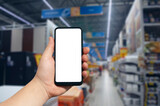 Fototapeta  - Online shopping using your phone. Mockup Smartphone with a white screen in hand close up on the background of shelving in a supermarket.