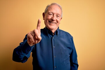 Wall Mural - Grey haired senior man wearing casual blue shirt standing over yellow background pointing finger up with successful idea. Exited and happy. Number one.