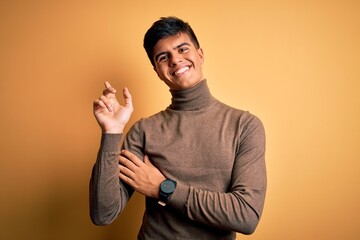 Canvas Print - Young handsome man wearing casual turtleneck sweater over isolated yellow background with a big smile on face, pointing with hand and finger to the side looking at the camera.