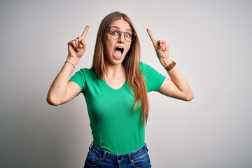 Wall Mural - Young beautiful redhead woman wearing casual green t-shirt and glasses over white background smiling amazed and surprised and pointing up with fingers and raised arms.