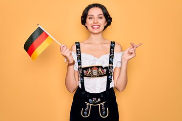 Wall Mural - Young patriotic german woman with blue eyes wearing octoberfest dress holding germany flag very happy pointing with hand and finger to the side