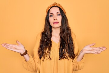 Wall Mural - Young beautiful woman wearing casual t-shirt and diadem standing over yellow background clueless and confused expression with arms and hands raised. Doubt concept.