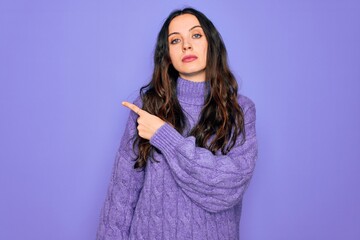 Wall Mural - Young beautiful woman wearing casual turtleneck sweater standing over purple background Pointing with hand finger to the side showing advertisement, serious and calm face