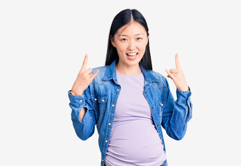 Sticker - Young beautiful chinese woman pregnant expecting baby shouting with crazy expression doing rock symbol with hands up. music star. heavy music concept.