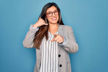 Sticker - Young hispanic business woman wearing glasses standing over blue isolated background smiling doing talking on the telephone gesture and pointing to you. Call me.