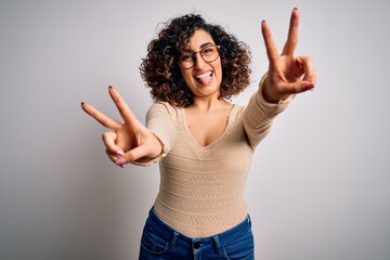 Wall Mural - Young beautiful curly arab woman wearing casual t-shirt and glasses over white background smiling with tongue out showing fingers of both hands doing victory sign. Number two.