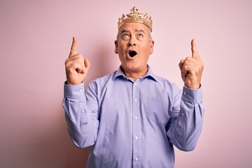 Wall Mural - Middle age hoary man wearing golden crown of king standing over isolated pink background amazed and surprised looking up and pointing with fingers and raised arms.