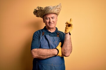 Wall Mural - Middle age hoary farmer man wearing apron and hat over isolated yellow background with a big smile on face, pointing with hand and finger to the side looking at the camera.