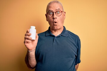 Middle age hoary man holding medicine pills standing over isolated yellow background scared in shock with a surprise face, afraid and excited with fear expression