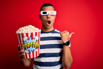 Young handsome african american man watching movie using 3d glasses eating popcorns Surprised pointing with hand finger to the side, open mouth amazed expression.