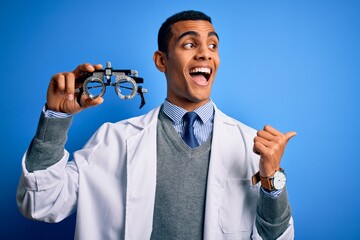 Wall Mural - Handsome african american optical man holding optometry glasses over blue background pointing and showing with thumb up to the side with happy face smiling