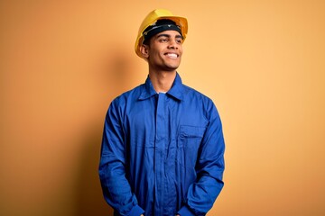 Wall Mural - Young handsome african american worker man wearing blue uniform and security helmet looking away to side with smile on face, natural expression. Laughing confident.