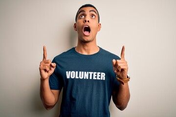 Wall Mural - Young handsome african american man volunteering wearing t-shirt with volunteer message amazed and surprised looking up and pointing with fingers and raised arms.