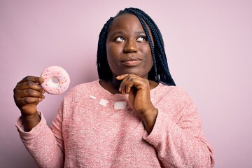 Sticker - Young african american plus size woman with braids eating doughnut over pink background serious face thinking about question, very confused idea