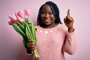 Sticker - Young african american plus size woman with braids holding bouquet of pink tulips flower pointing finger up with successful idea. Exited and happy. Number one.