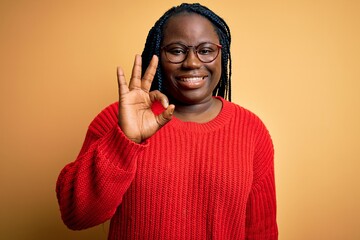 Poster - African american plus size woman with braids wearing casual sweater over yellow background smiling positive doing ok sign with hand and fingers. Successful expression.