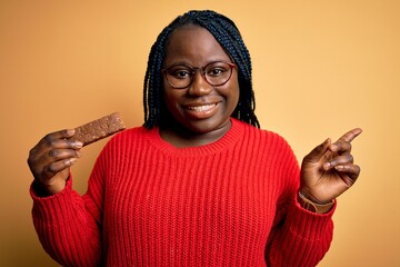 Sticker - Young african american plus size woman with braids eating healthy protein bar very happy pointing with hand and finger to the side