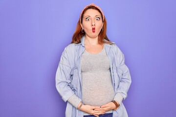 Poster - Young beautiful redhead pregnant woman expecting baby over isolated purple background making fish face with lips, crazy and comical gesture. Funny expression.