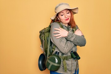 Poster - Young redhead backpacker woman hiking wearing backpack and hat over yellow background Hugging oneself happy and positive, smiling confident. Self love and self care