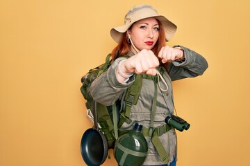 Wall Mural - Young redhead backpacker woman hiking wearing backpack and hat over yellow background Punching fist to fight, aggressive and angry attack, threat and violence