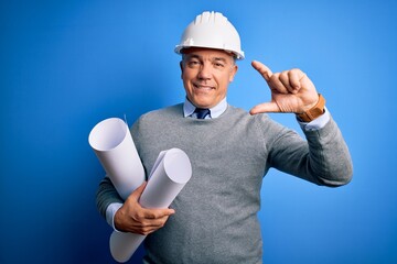 Sticker - Middle age handsome grey-haired architect man wearing safety helmet holding blueprints smiling and confident gesturing with hand doing small size sign with fingers looking and the camera. Measure