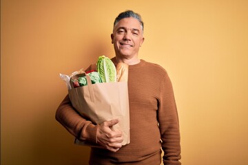 Poster - Middle age handsome grey-haired man holding paper bag with food over yellow background with a happy face standing and smiling with a confident smile showing teeth