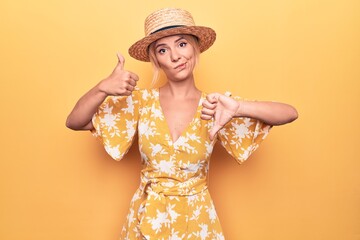 Wall Mural - Beautiful blonde woman on vacation wearing summer hat and dress over yellow background Doing thumbs up and down, disagreement and agreement expression. Crazy conflict