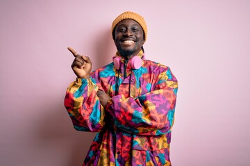 Young handsome african american man wearing colorful coat and cap over pink background with a big smile on face, pointing with hand and finger to the side looking at the camera.