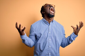 Poster - Young handsome african american man wearing shirt and glasses over yellow background crazy and mad shouting and yelling with aggressive expression and arms raised. Frustration concept.