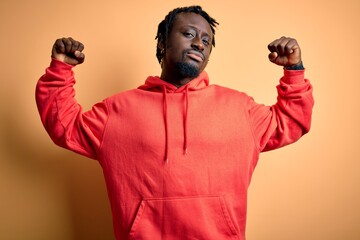 Poster - Young african american sporty man wearing sweatshirt with hoodie over yellow background showing arms muscles smiling proud. Fitness concept.