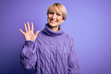 Sticker - Young blonde woman with short hair wearing winter turtleneck sweater over purple background showing and pointing up with fingers number five while smiling confident and happy.