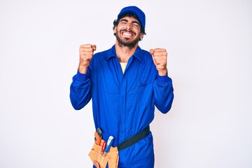 Handsome young man with curly hair and bear weaing handyman uniform excited for success with arms raised and eyes closed celebrating victory smiling. winner concept.