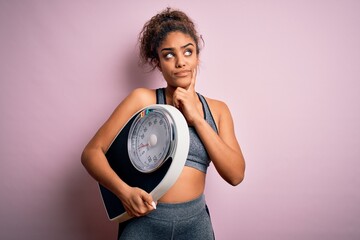 Wall Mural - Young african american sporty girl holding weighing machine over isolated pink background serious face thinking about question, very confused idea