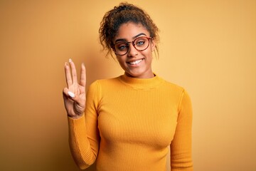 Sticker - Young beautiful african american girl wearing sweater and glasses over yellow background showing and pointing up with fingers number three while smiling confident and happy.