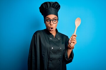 Canvas Print - Young african american chef girl wearing cooker uniform and hat holding wooden spoon tool scared in shock with a surprise face, afraid and excited with fear expression