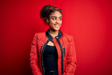 Canvas Print - Beautiful african american girl wearing red jacket and sunglasses over isolated background looking away to side with smile on face, natural expression. Laughing confident.