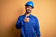 © Krakenimages.com - Mechanic man with beard wearing blue uniform and safety helmet over yellow background doing happy thumbs up gesture with hand. Approving expression looking at the camera showing success.