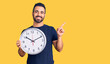 © Krakenimages.com - Young hispanic man holding big clock smiling happy pointing with hand and finger to the side