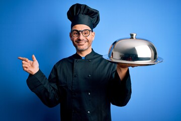 Young handsome chef man wearing uniform and hat holding waiter tray with dome very happy pointing with hand and finger to the side