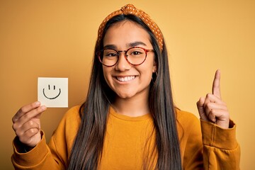 Poster - Asian woman wearing glasses holding reminder with smile emoji draw over yellow background surprised with an idea or question pointing finger with happy face, number one