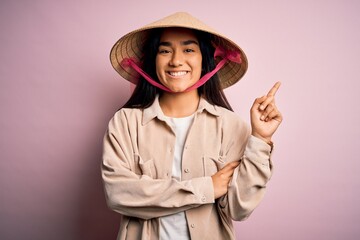 Canvas Print - Young beautiful woman wearing traditional conical asian hat over isolated pink background with a big smile on face, pointing with hand and finger to the side looking at the camera.