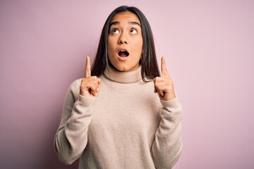 Canvas Print - Young beautiful asian woman wearing casual turtleneck sweater over pink background amazed and surprised looking up and pointing with fingers and raised arms.